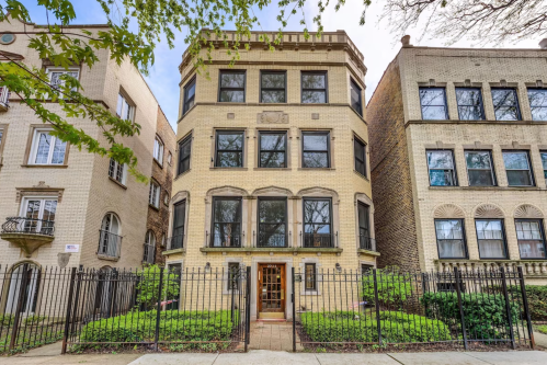 A three-story brick building with large windows, surrounded by greenery and a black iron fence.