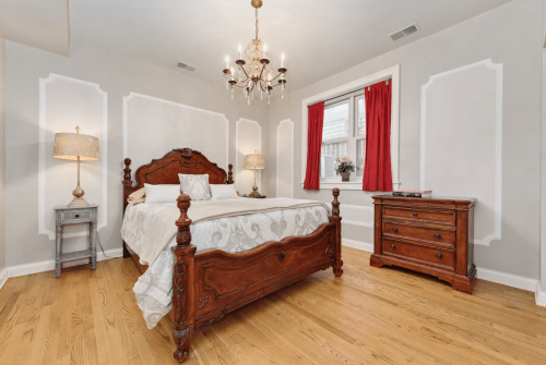 Cozy bedroom featuring a wooden bed, chandelier, red curtains, and elegant decor on light walls.