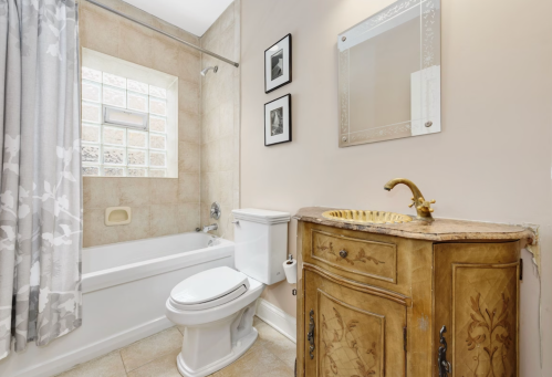 A bright bathroom featuring a bathtub, toilet, decorative mirror, and a vintage wooden cabinet with floral details.