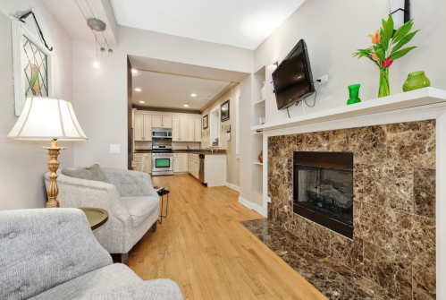 Cozy living room with a fireplace, modern kitchen in the background, and stylish decor including a lamp and plants.