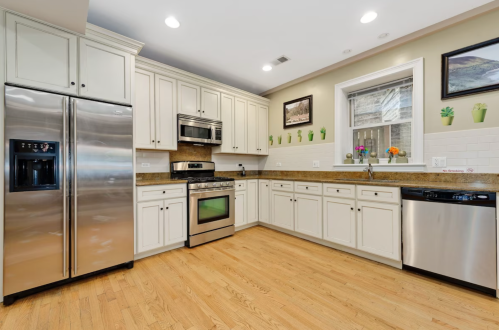 Modern kitchen with stainless steel appliances, light cabinets, and wooden flooring, featuring potted plants on the walls.