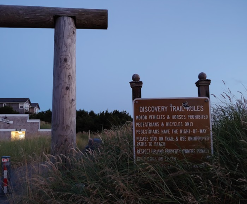 Sign at the Discovery Trail entrance, stating rules for pedestrians and bicycles, surrounded by tall grass at dusk.