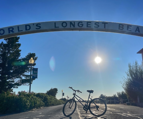 A bicycle rests under an archway that reads "World's Longest Beach," with a bright sun in the background.