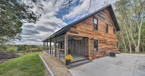 A rustic wooden house with a porch, surrounded by greenery and a cloudy sky in the background.