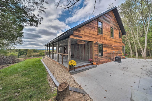 A rustic wooden house with a porch, surrounded by greenery and a cloudy sky.