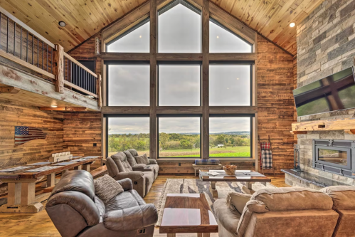 Spacious rustic living room with large windows, wooden walls, and cozy seating, overlooking a scenic landscape.
