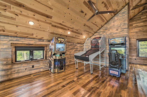 A cozy attic room with wooden walls, featuring a bed and vintage arcade games near large windows.
