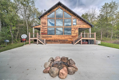 A modern wooden cabin with large windows, surrounded by trees, and a stone fire pit in the foreground.
