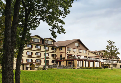 A large, multi-story lodge with balconies, surrounded by trees and green grass under a cloudy sky.