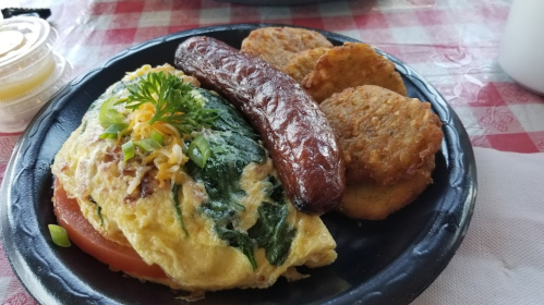 A plate with a spinach omelet, sausage, and golden-brown potato cakes on a checkered tablecloth.