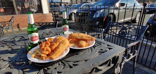 Two plates of fried fish, shrimp, and fries on a table with two bottles of beer, parked cars in the background.
