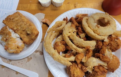 A plate of fried food including onion rings, chicken pieces, and a side of fried fish on a paper plate.