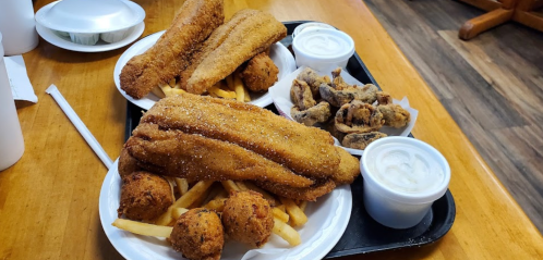 Two trays of fried fish fillets, hushpuppies, and fries, served with dipping sauces on a wooden table.