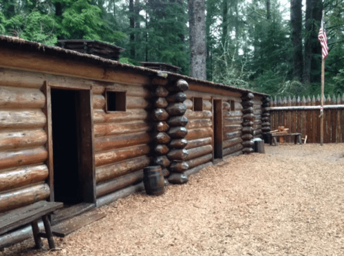 Log cabin structure surrounded by trees, featuring wooden walls, a flag, and a gravel path.