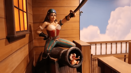A pirate woman poses confidently on a cannon, with a small figure peeking out from inside the cannon.