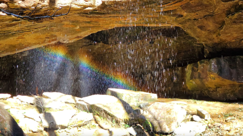 A cave with a waterfall creating a rainbow effect, surrounded by rocks and natural light.