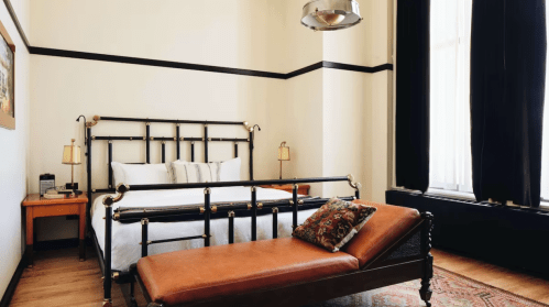 A stylish bedroom featuring a metal bed frame, a chaise lounge, and large windows with dark curtains.