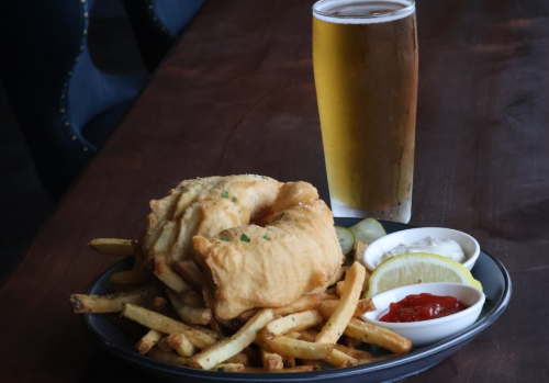 A plate of golden fried fish and fries, served with lemon wedges and dipping sauces, alongside a tall glass of beer.