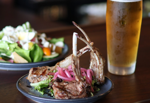 A plate with grilled lamb chops, pickled onions, and greens, alongside a salad and a glass of beer.