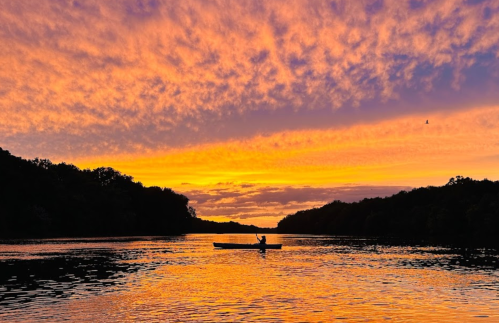 A person kayaking on a calm river at sunset, with vibrant orange and purple clouds reflecting on the water.