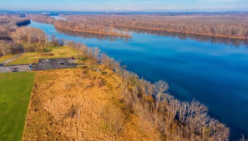 Aerial view of a calm river bordered by bare trees and grassy areas, with a parking lot visible nearby.