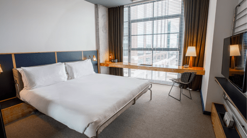 Modern hotel room with a large bed, minimalist decor, a desk, and large windows letting in natural light.