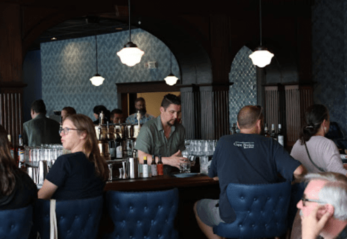 A bar scene with patrons seated at a counter, a bartender preparing drinks, and a stylish interior.