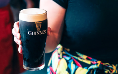 A person holding a pint of Guinness beer with a creamy head, wearing a black shirt and colorful patterned pants.