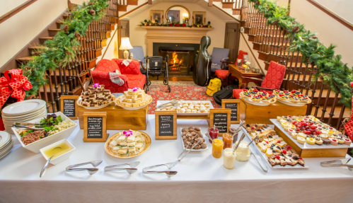 A festive dessert table with various treats, surrounded by holiday decorations and a cozy fireplace in the background.