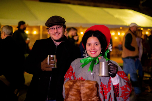 A man and woman in festive attire hold mugs, smiling at a lively outdoor event with string lights in the background.