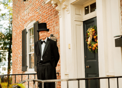 A man in a top hat and tuxedo stands by a door adorned with a festive wreath, in front of a brick building.