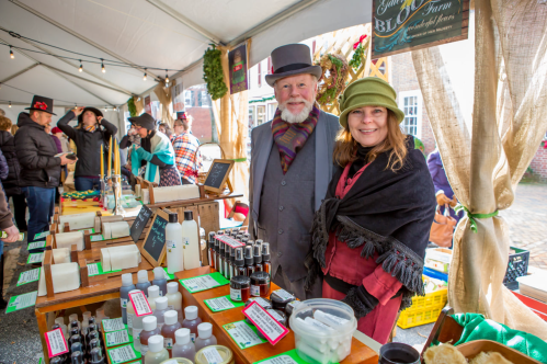 A man and woman in vintage attire stand behind a table of handmade soaps and skincare products at a market.