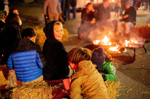 A group of children sitting on hay bales near a fire, enjoying a festive evening outdoors.