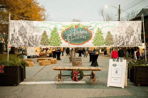 A festive banner for a Dickens Christmas event, displayed over a street with hay bales and tables set up.