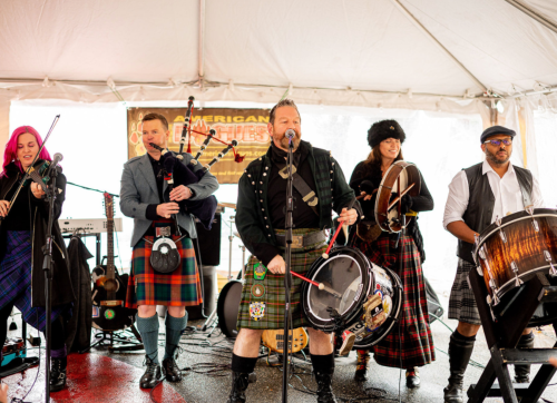 A lively band in kilts performs under a tent, featuring bagpipes, drums, and various instruments.