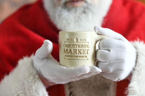 A close-up of Santa's hands holding a mug that reads "Christkindl Market 2023." Santa is wearing a red coat and white gloves.