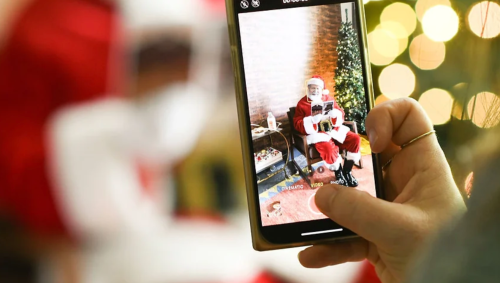 A person takes a photo of Santa Claus sitting in a chair, surrounded by Christmas decorations and lights.