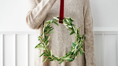 A person holding a circular olive leaf wreath with a red ribbon against a light background.
