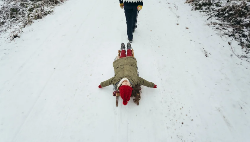A child in winter clothing lies on a snowy path, while an adult walks nearby.