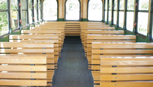 Interior of a bus with wooden benches arranged in rows, featuring large windows and a clear aisle.