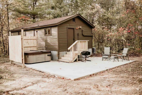 A small brown cabin with a deck, hot tub, and outdoor seating in a wooded area.