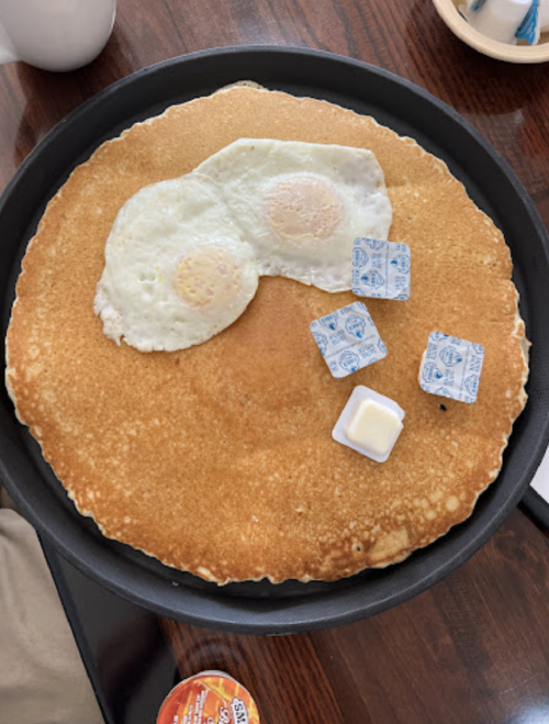 A large pancake with two fried eggs resembling glasses, accompanied by butter and syrup packets on a black plate.