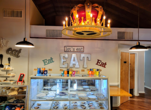 A cozy café interior featuring a crown chandelier, colorful "EAT" signage, and a display case with various desserts.