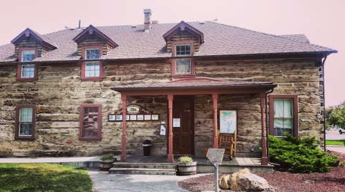 Historic stone building with a porch, multiple windows, and a signboard in front, surrounded by greenery.