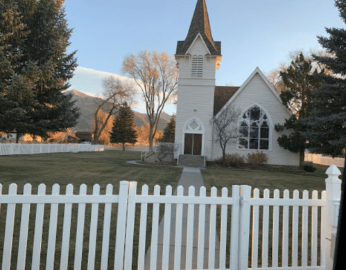 A white church with a steeple, surrounded by trees and a white picket fence, set against a mountain backdrop.