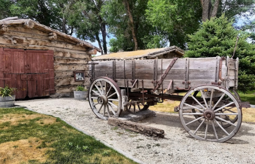 A weathered wooden wagon sits beside a rustic log cabin, surrounded by greenery and gravel pathways.
