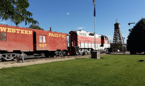 A red Western Pacific train car and locomotive beside a wooden water tower on a sunny day. Green grass in the foreground.
