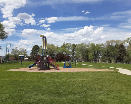 A playground with colorful equipment on a grassy field, surrounded by trees and a blue sky with clouds.