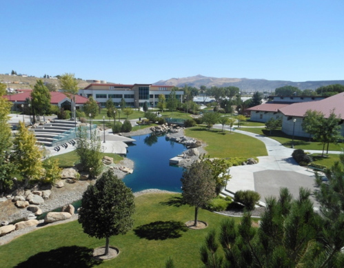A serene park with a pond, walking paths, trees, and buildings in the background under a clear blue sky.