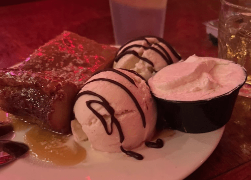 A plate with a slice of cake, two scoops of ice cream drizzled with chocolate, and a small cup of whipped cream.